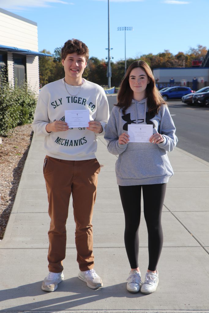 Two teenagers stand outside a school with certificates.