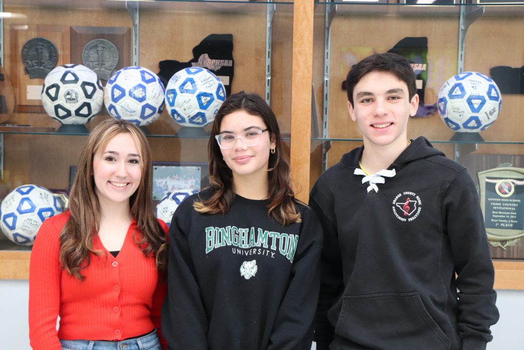 Three teenagers stand together in front of a display case with signed soccer balls and awards. 