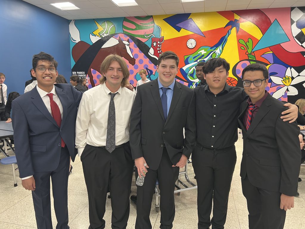 A group of teenagers stand together with arms around each other's shoulders in a cafeteria in front of a colorful mural. 