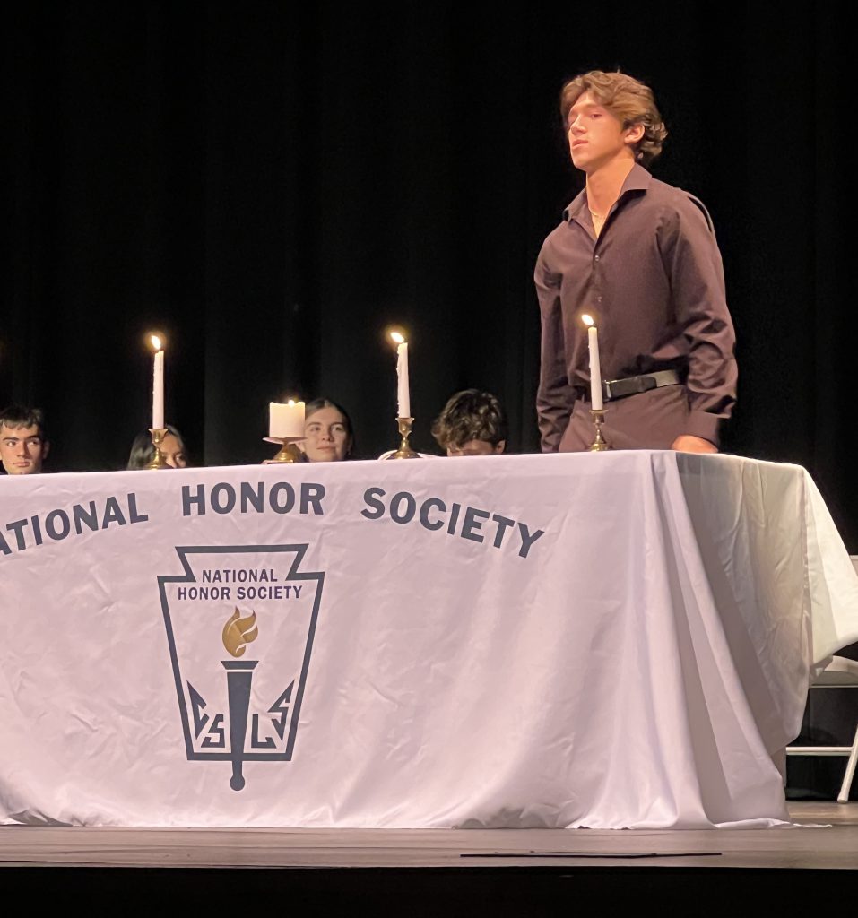 A teenager stands in front of a table with candles on it that says "National Honor Society" on a stage.