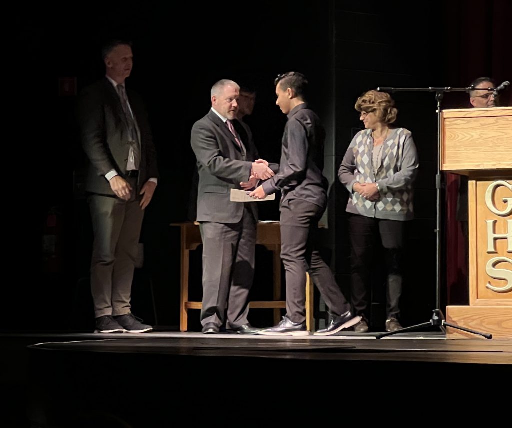 A teenager shakes the hand of a man wearing a suit on a stage next to a podium that says "GHS."