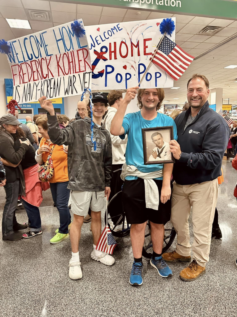 Teenagers and an adult hold signs and a photograph of a U.S. Navy veteran at an airport. The signs say "Welcome Home Frederick Kohler U.S. Navy-Korean War Vet" and "Welcome Home Pop Pop."