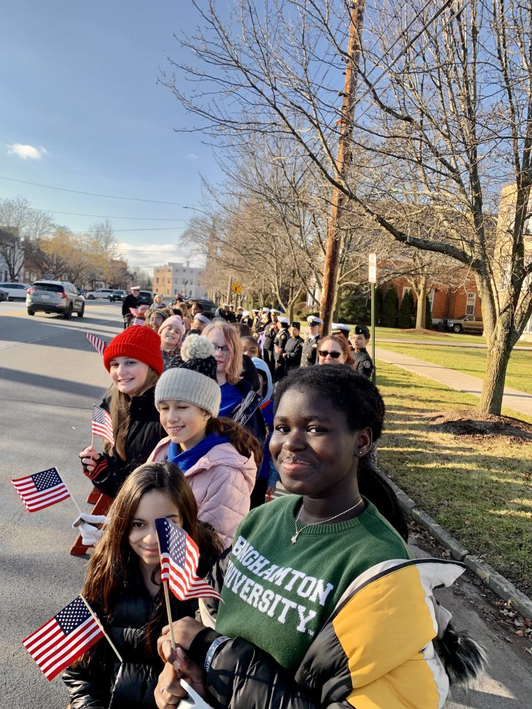 Middle school students stand in front of the Goshen School District Main Office Building holding small American flags.