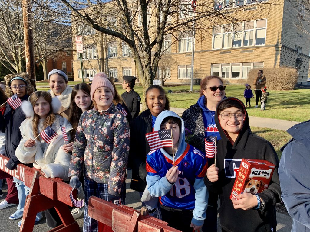 Veterans and middle school students stand in front of the Goshen School District Main Office Building holding small American flags and a box of Milk-Bone dog biscuits.