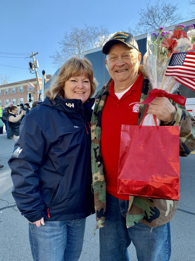 A woman and a man in a Vietnam Veteran baseball cap stand together, with the man holding a gift bag, flowers and small American flags.
