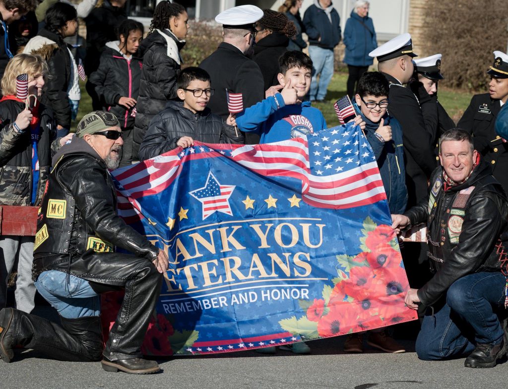 Veterans, bikers and middle school students stand in front of the Goshen School District Main Office Building holding small American flags and a banner that says "Thank You Veterans."