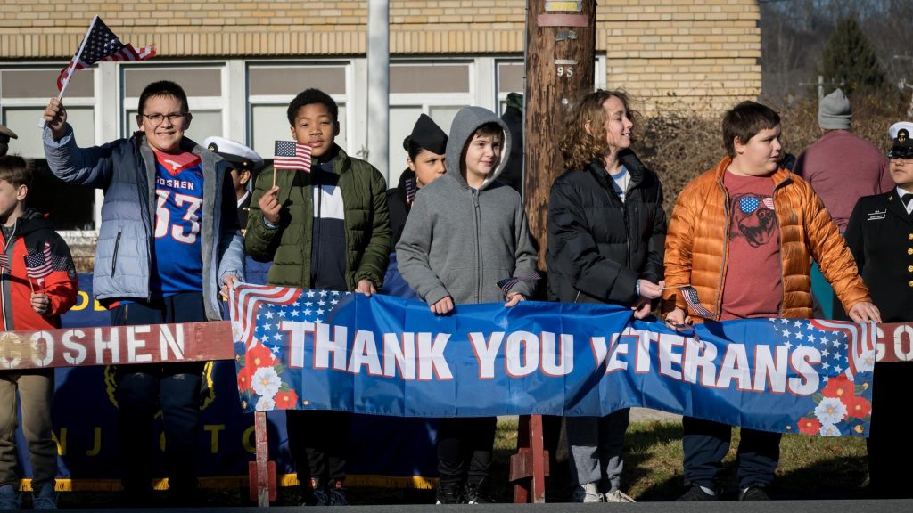 Middle school students stand in front of the Goshen School District Main Office Building holding small American flags and a banner that says "Thank You Veterans."