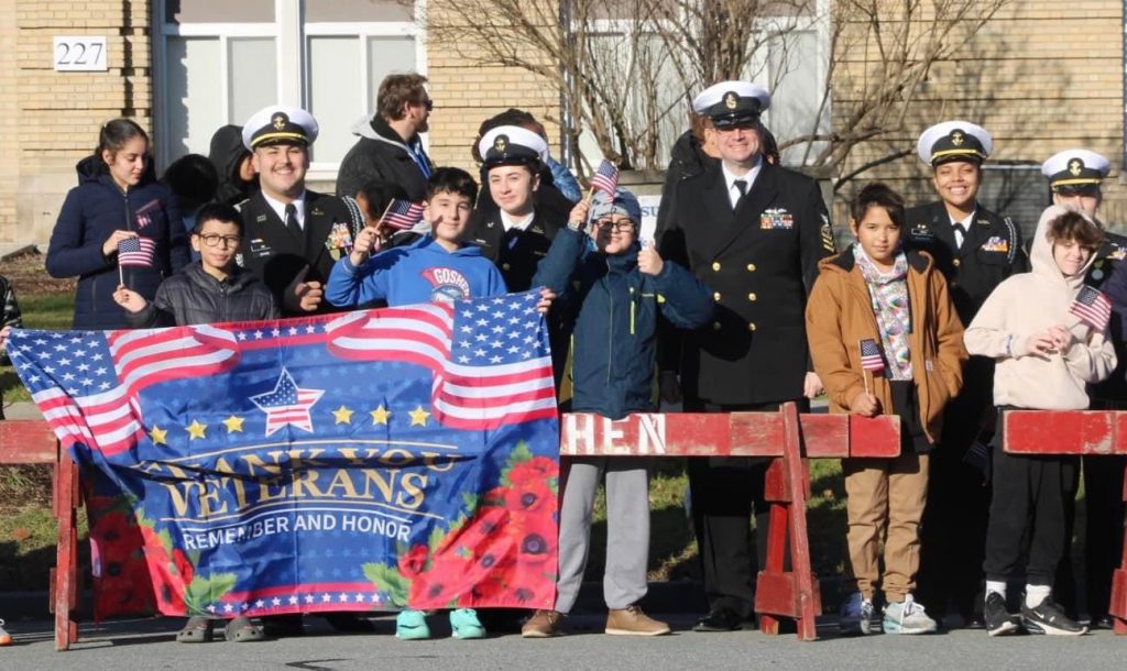 Veterans and middle school students stand in front of the Goshen School District Main Office Building holding small American flags and a banner that says "Thank You Veterans."