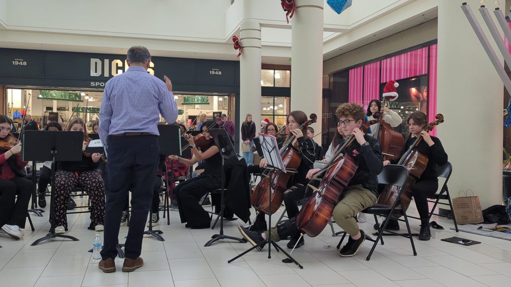 A middle school orchestra performs music in front of a Dick's Sporting Goods in a mall.
