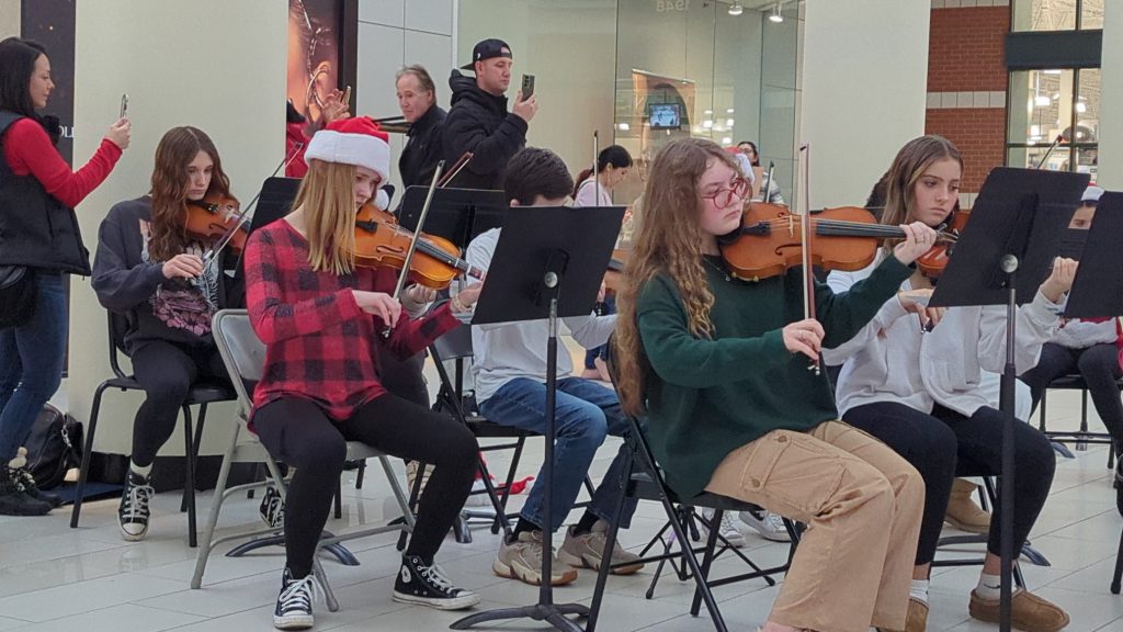 Middle school students play violin in Santa hats at the mall while passersby film on their smart phones. 