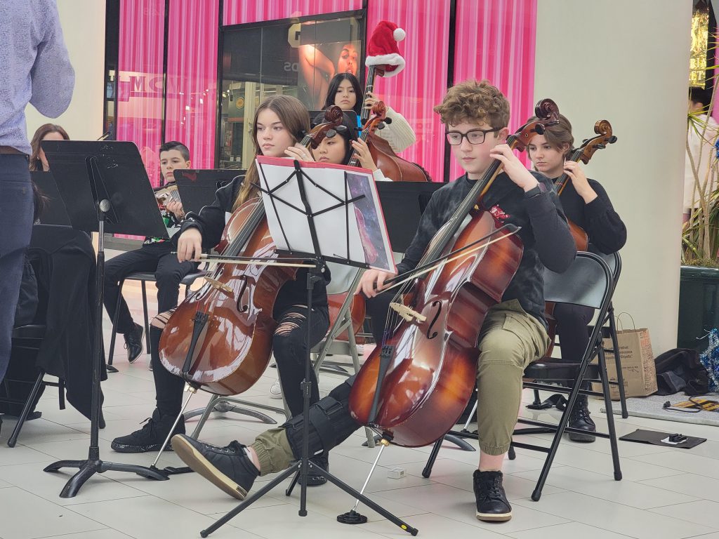 Middle school students play large orchestra instruments in front of a Victoria's Secret in a mall. 