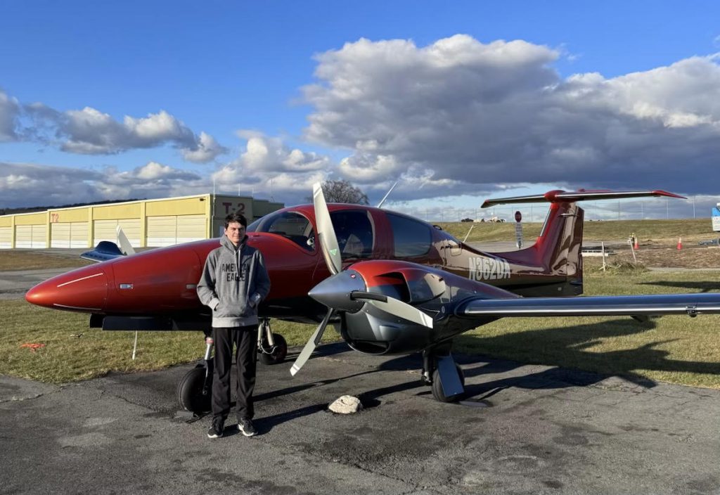 A teenager stands in front of a small red plane with a vast expanse of sky behind him.