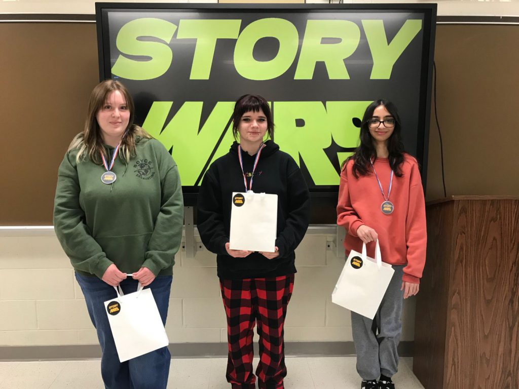 Three high school students stand in front of a large green and black screen that says "STORY WARS," holding white paper bags and wearing medals. 