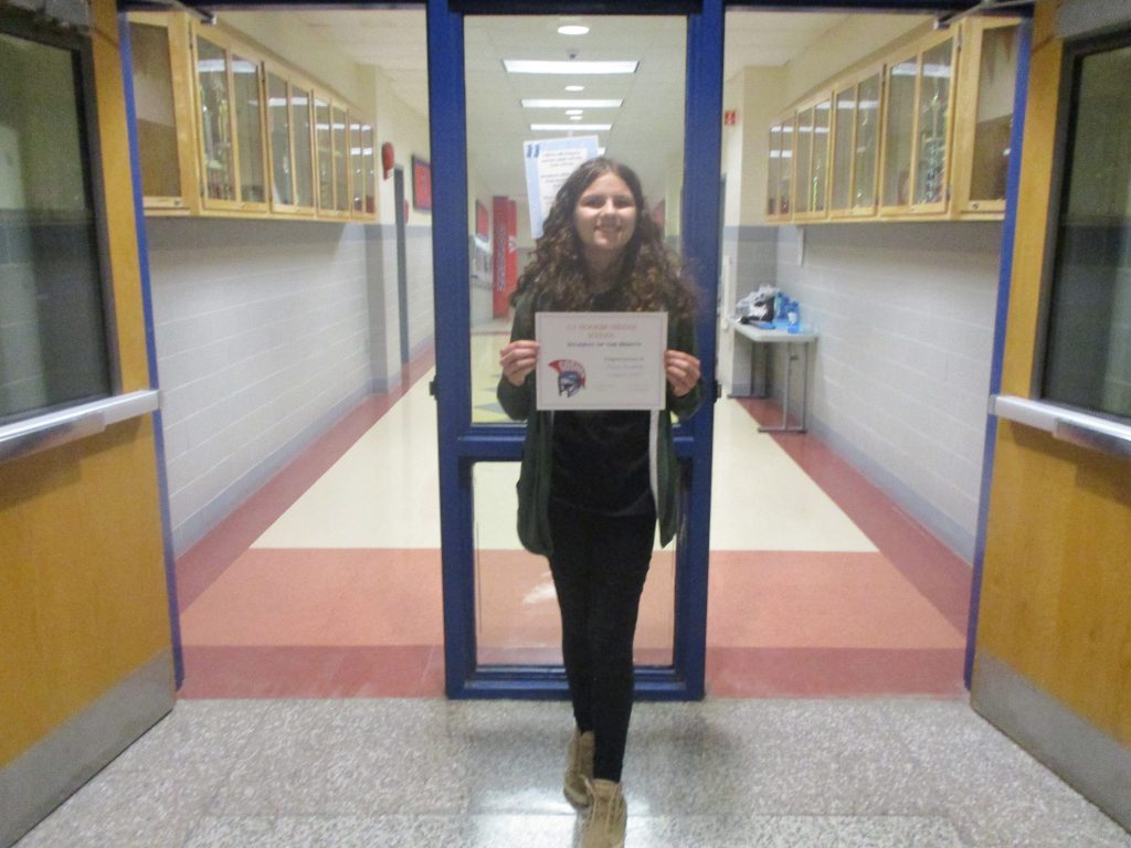 A middle school student holds a certificate in front of her while smiling in a school hallway. 