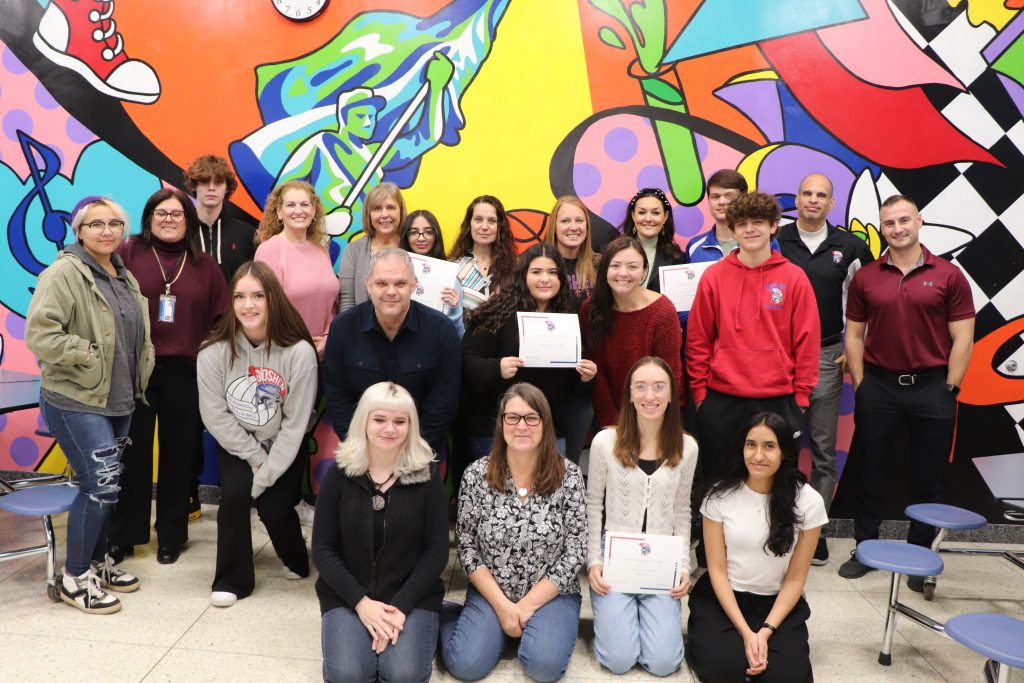 All of Goshen High School's Q1 Goshen Greats for the 2023-24 school year smiled for a photo with the teachers who nominated them in front of a colorful mural in the cafeteria.