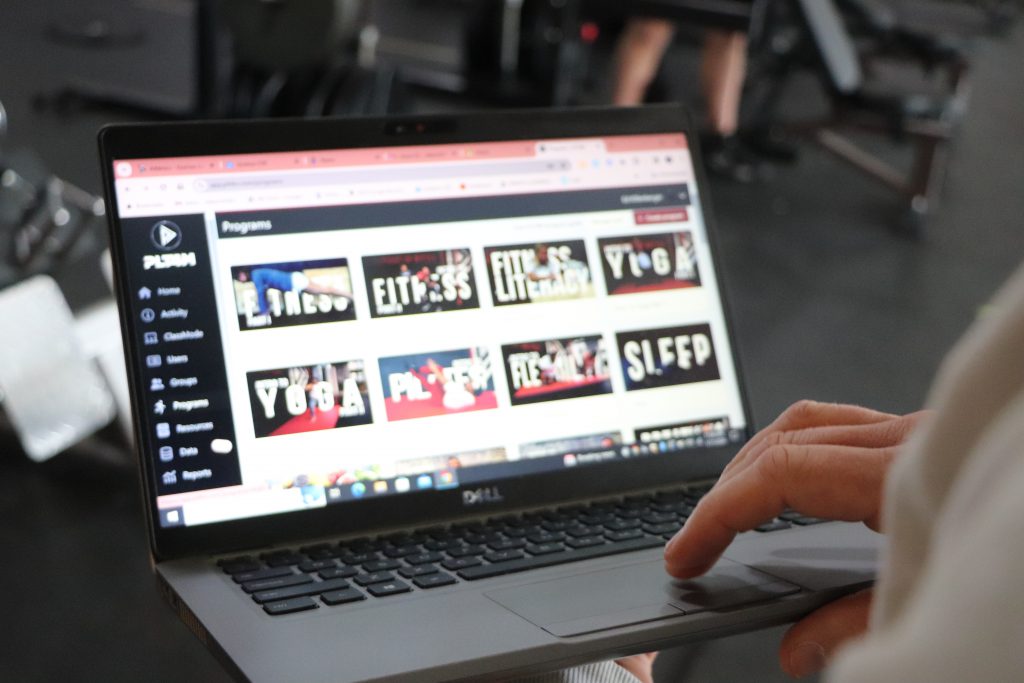 A man navigates a web page with instructional physical education videos on a laptop in a weight room.
