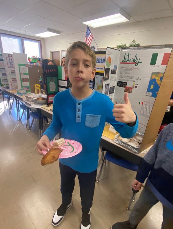 An elementary school student holds a paper plate with food on it in one hand and gives a thumbs up with the other hand in front of informative trifolds depicting various cultures in the Western Hemisphere in a classroom.