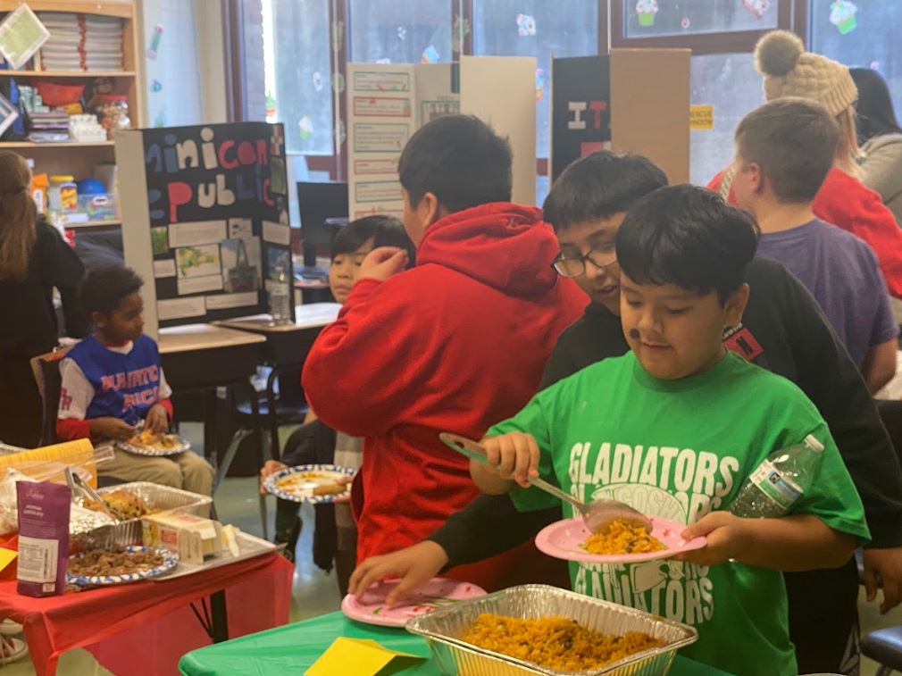 Elementary school students load up food on paper plates in a classroom surrounded by informative tri-folds for various cultures in the Western Hemisphere.