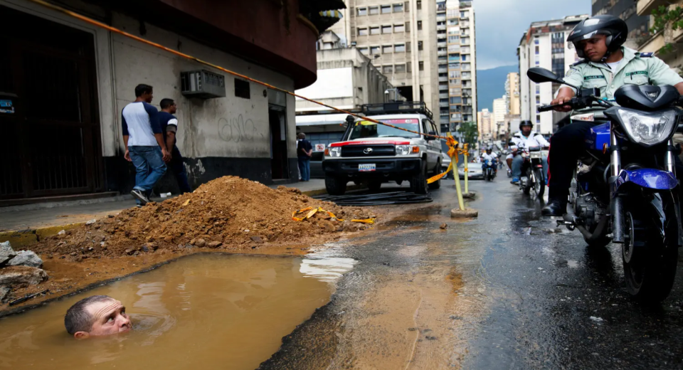 A man's head sticks up out of a brown puddle in the street. Tape ropes off the area, next to a large dirt pile and truck, while people on motorcycles drive by.