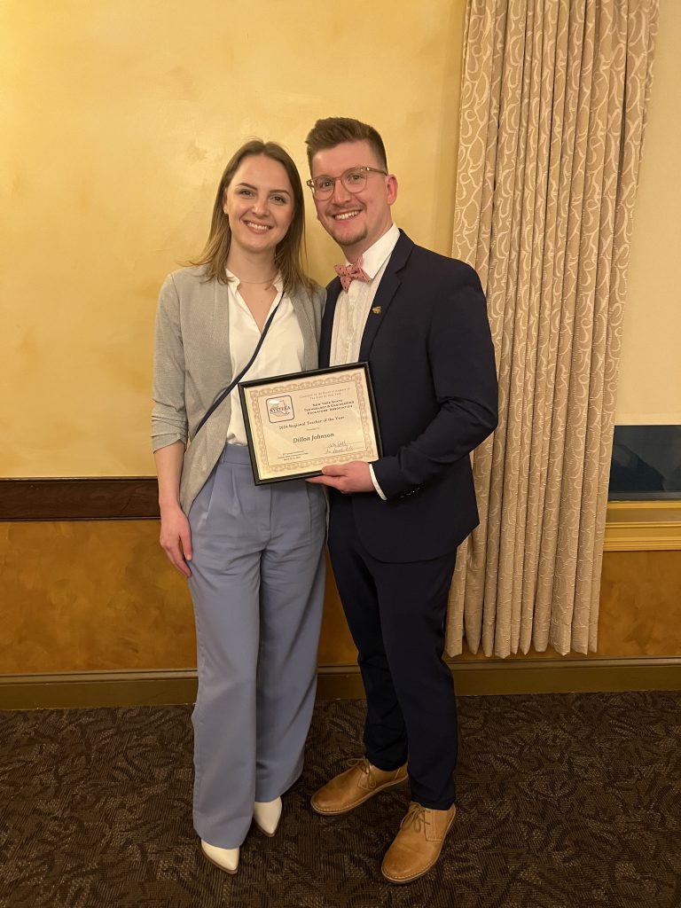 Two people stand together smiling in front of a wall and curtain. One is holding an award.,