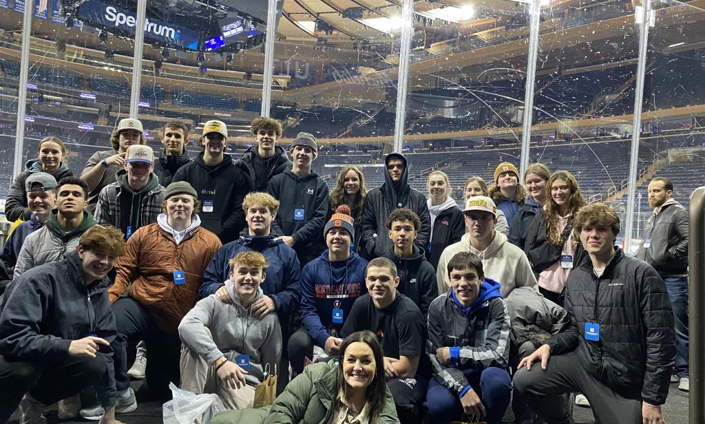 A group of high school students smile together with their teacher next to an ice rink at Madison Square Garden.