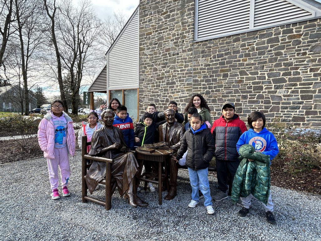 A group of intermediate school students stand next to a sculpture of Franklin D. and Eleanor Roosevelt outside a historic home.