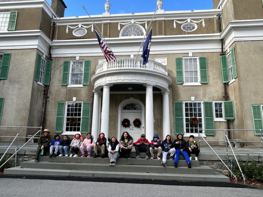 A group of intermediate school students sits on the steps of a historic home.