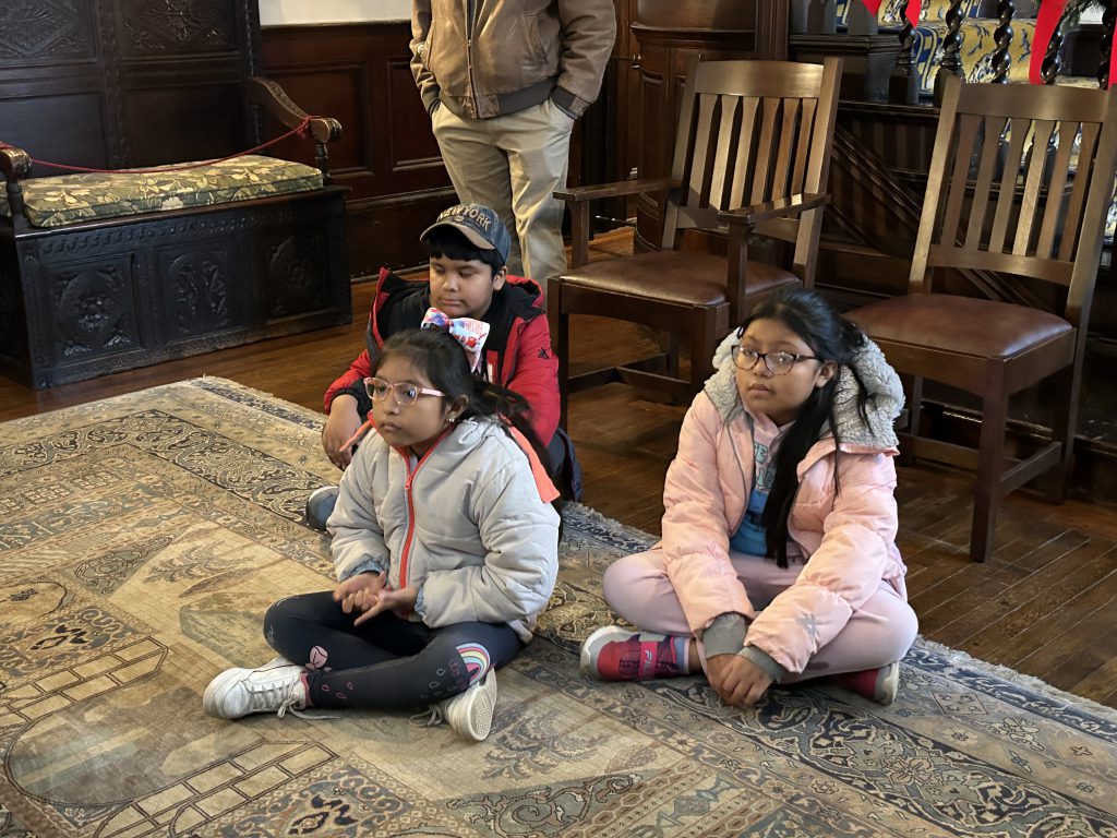 A group of intermediate school students sit on the floor of a historic home next to wooden chairs, a bench.