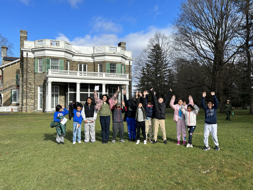 A group of intermediate school students smile and raise their arms together in front of a historic home.