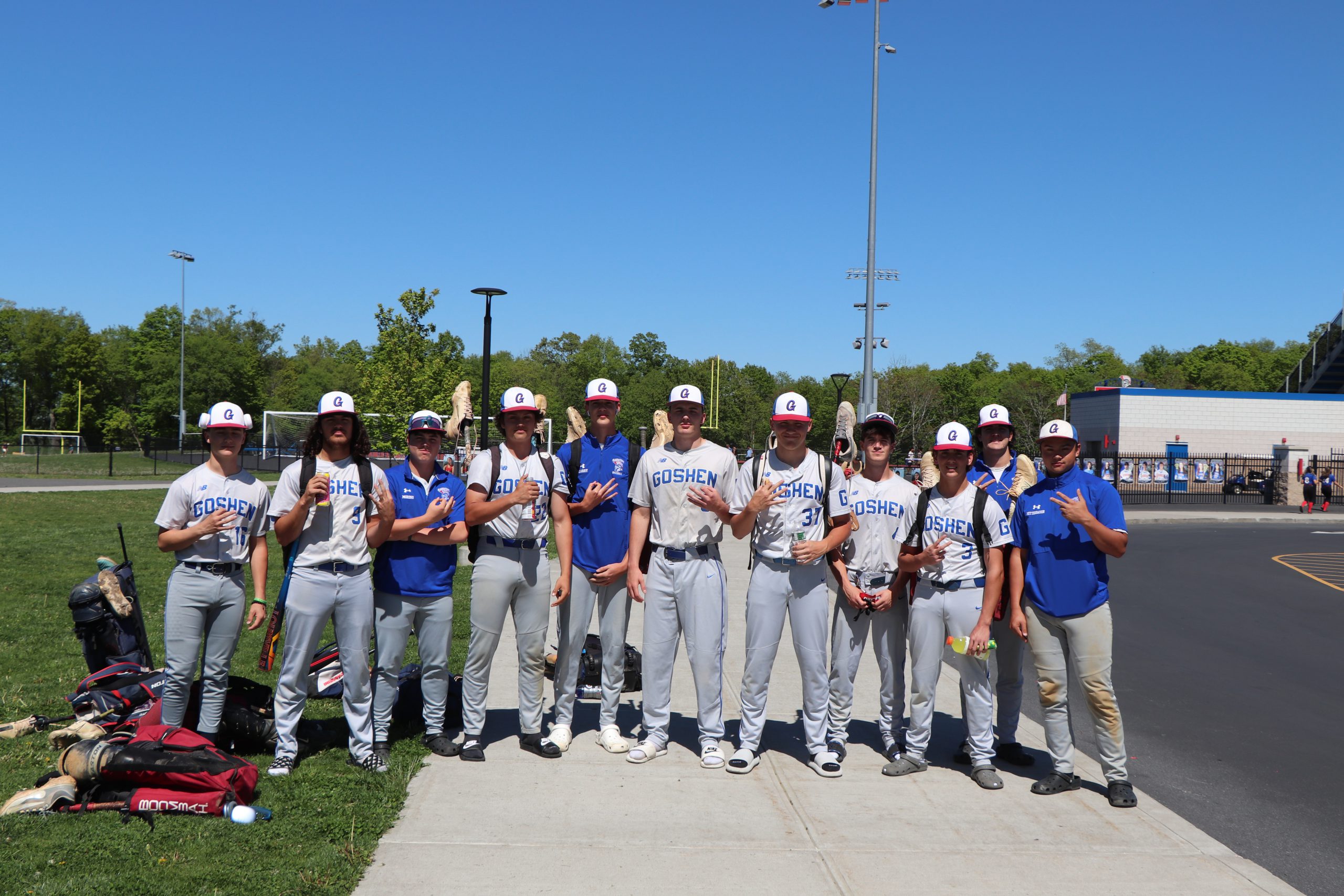 GHS Baseball team throws up hand signals standing together in uniform with gear to their left outside the high school.
