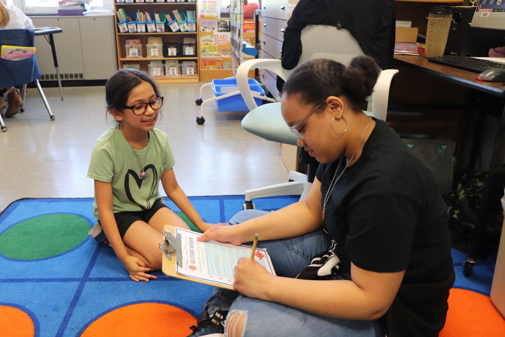 A high school student and an elementary school student sit on a carpet in a classroom talking, while the high school student takes notes on a clipboard.