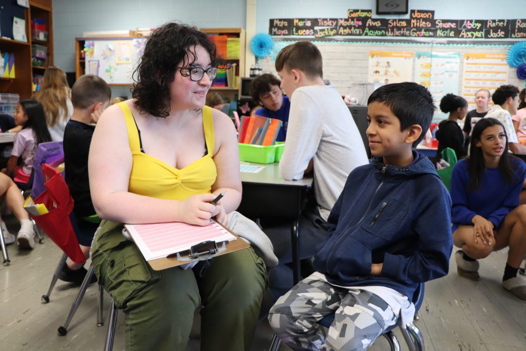 A high school student and an elementary school student sit in chairs in an elementary school classroom smiling at each other.