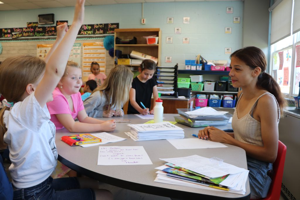 A high school student sits on one side of a u-shaped table with two elementary school students across from her. One holds her hands up in the air excitedly. Other students are talking to each other in the background.