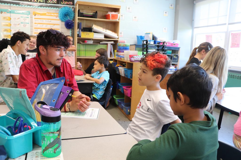 A high school student sits with two elementary school students at a desk in an elementary school classroom taking notes on a piece of paper.