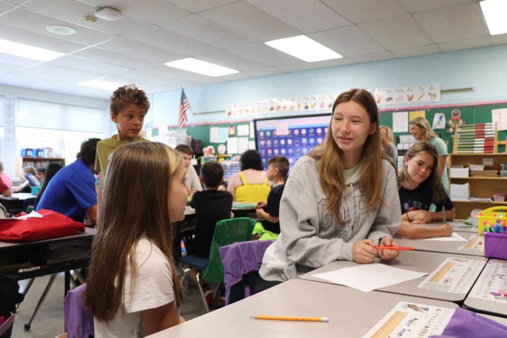 A high school student and elementary school student sit next to each other at a desk talking.