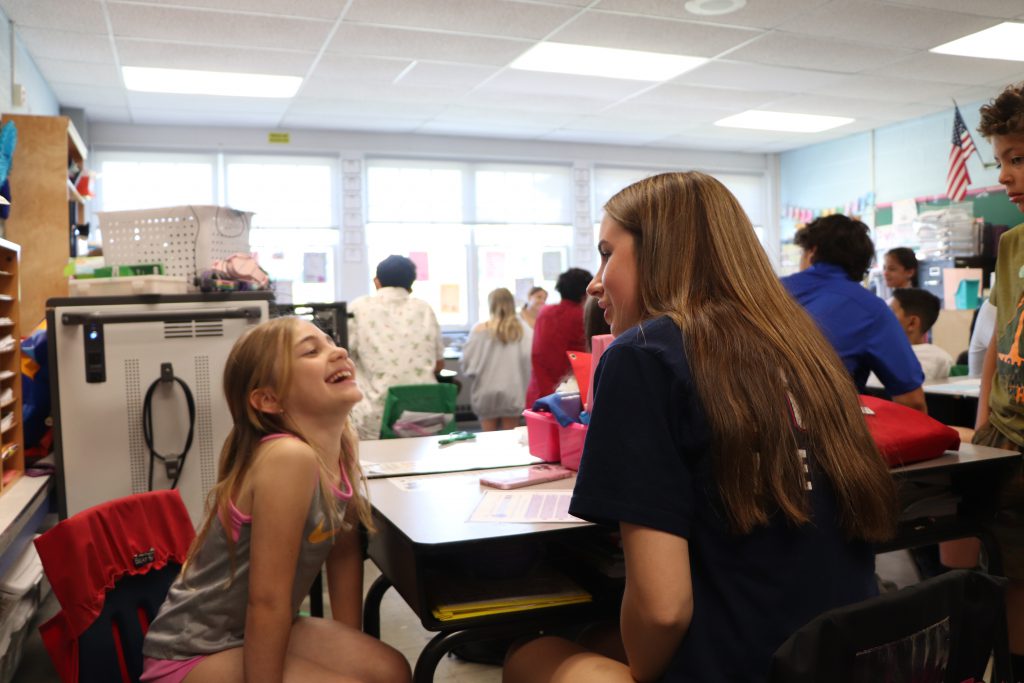 An elementary school student smiles with her shoulders shrugged and head leaning back talking to a high school student.