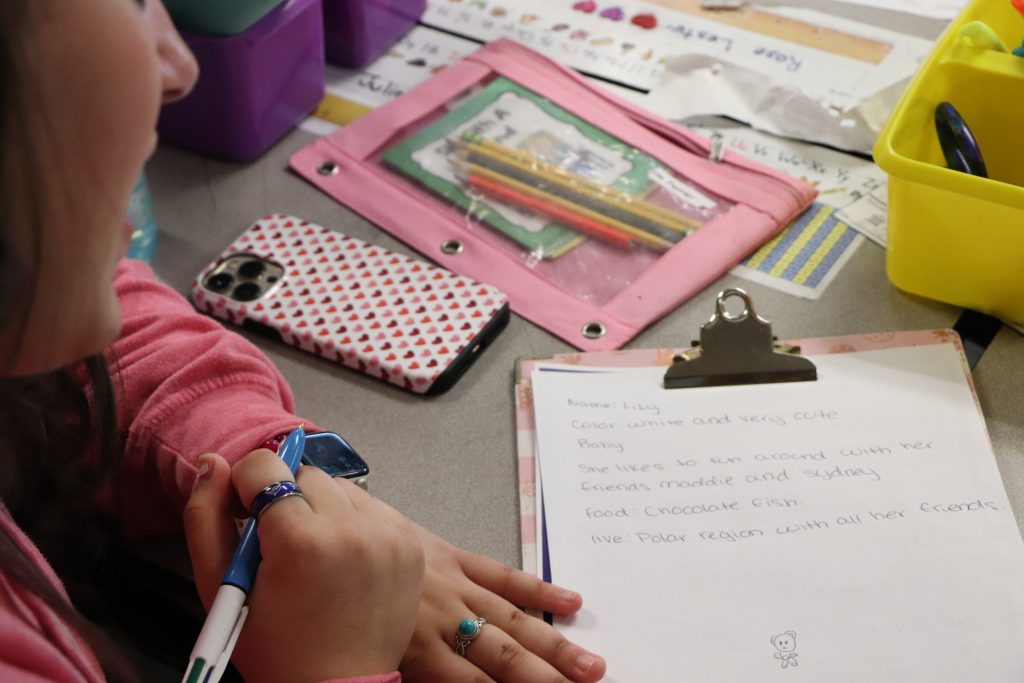 A clipboard with notes sits on a desk next to a pencil case and a cell phone with a high school student's hands crossed holding a pencil next to it.