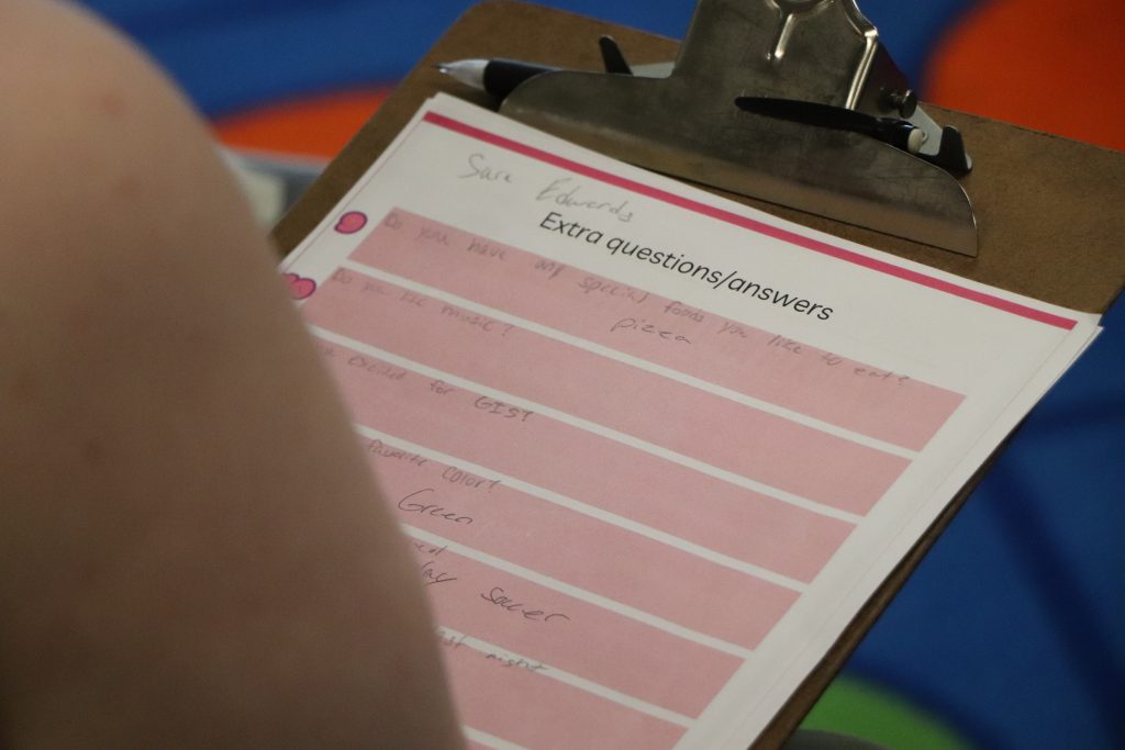 A worksheet on a clipboard titled "Extra questions/answers" has multiple pink spaces where text reads, "Do you have any special foods you like to eat? Pizza," "Do you like music?" and "Are you excited for GIS?" are written.