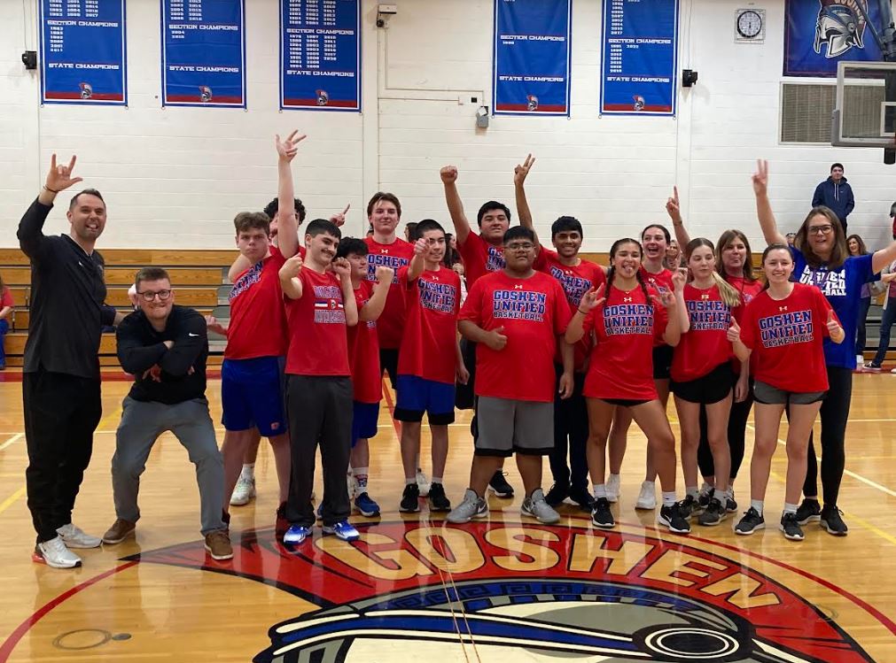 High school students wearing red T-shirts that say "Goshen Unified Basketball" raise their arms and pose with peace signs and hand gestures in a school gymnasium.