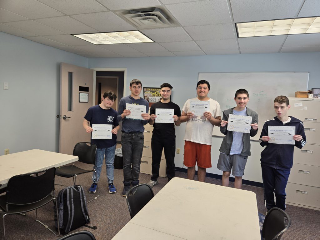 A group of high school students stand together smiling holding certificates in a classroom.