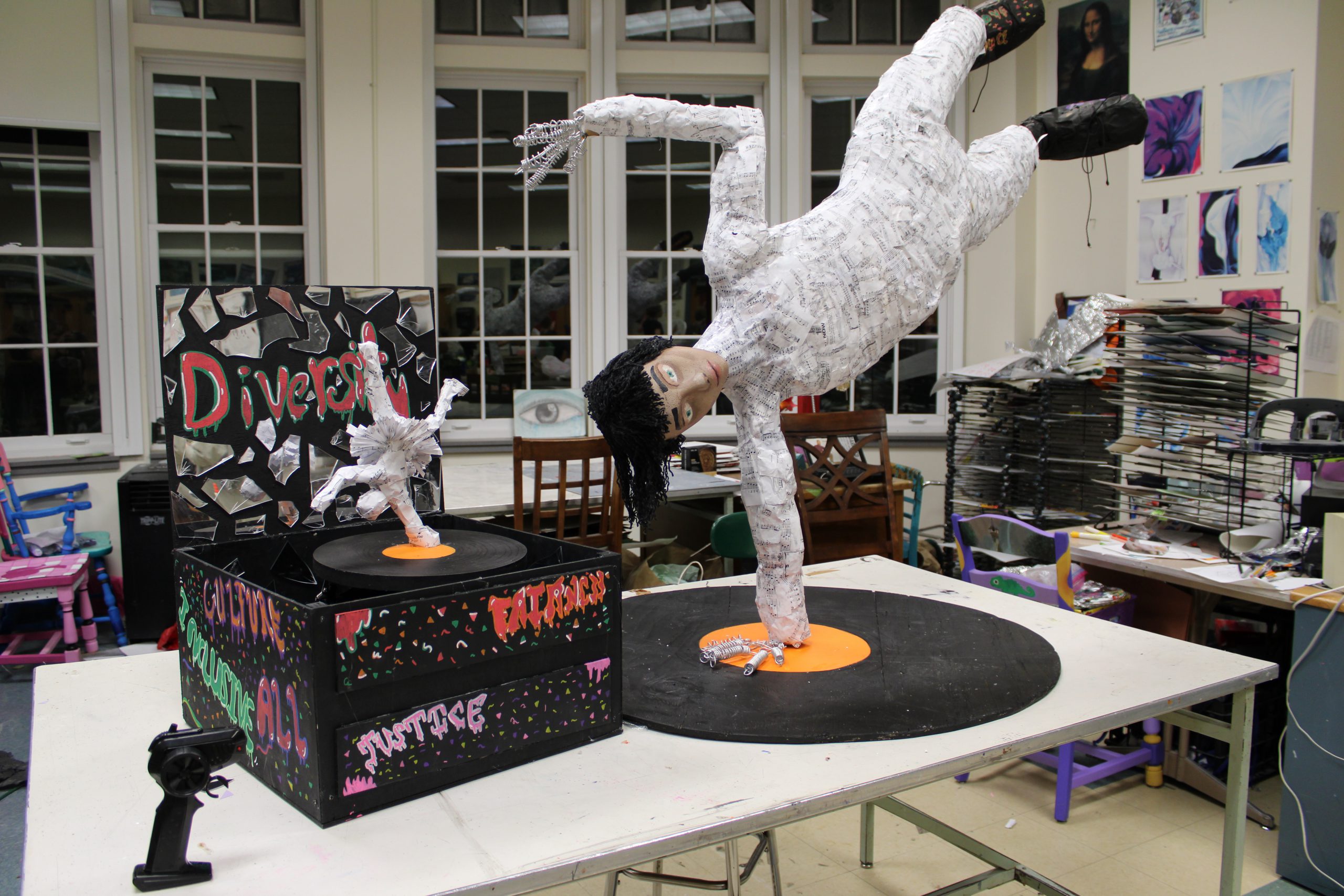 A papier mache sculpture of a person breakdancing on top of a black and orange vinyl record stands next to a miniature version of a person with no face in ballerina attire breakdancing on a record inside a black music box with glued pieces of mirrors and the text "Diversity" written across the top.