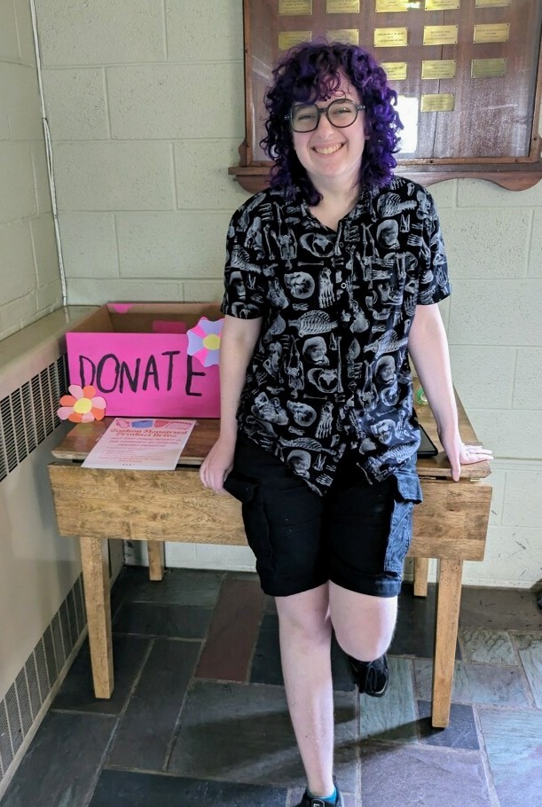 A high school student smiles standing in front of a desk and a pink box labeled "Donate."