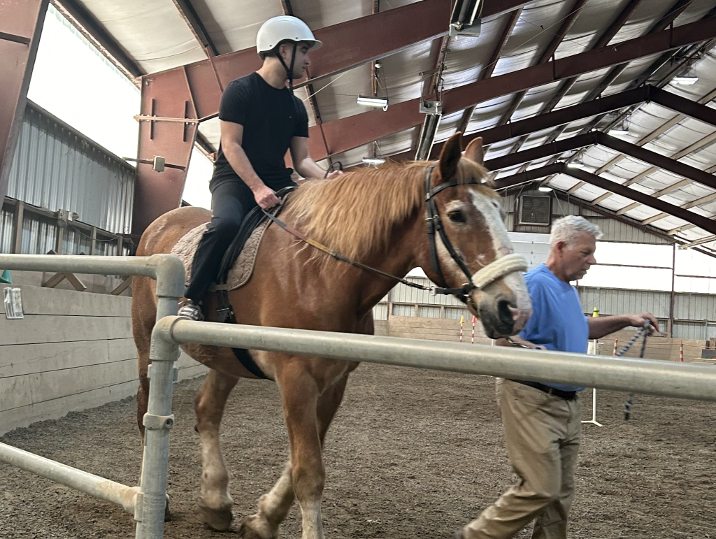 A high school student wearing a white helmet rides a brown horse in an indoor corral with an adult standing next to him.