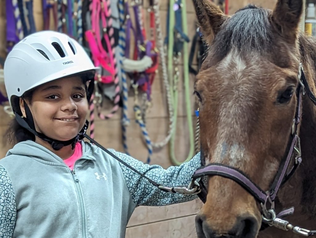 An intermediate school student smiles wearing a white helmet and petting a brown horse in a stable with colorful harnesses hanging on the wall behind them.