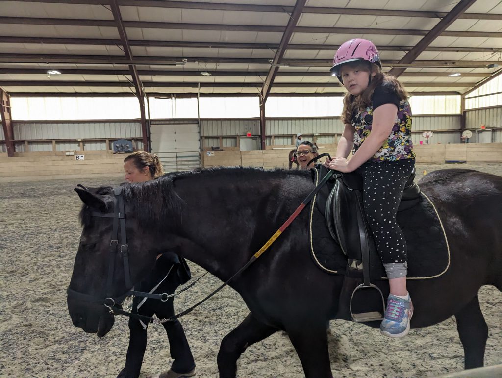 AN intermediate school student in a pink helmet sits atop a black horse in an indoor corral with adults standing next to them.