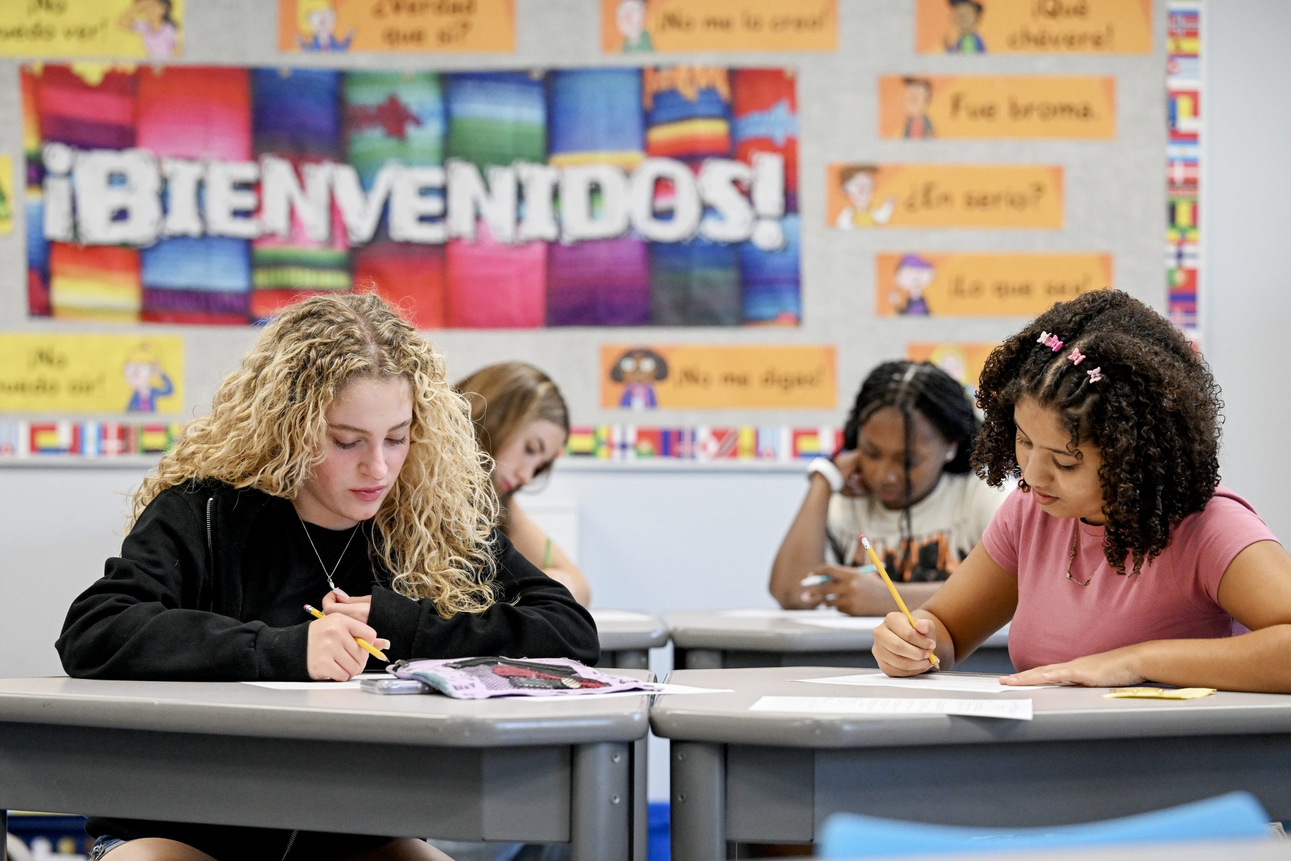 Teenagers sit at desks in a classroom working with a sign behind them that says bienvenidos.