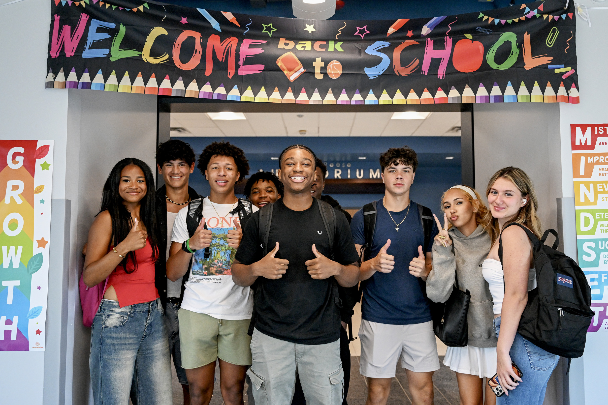 A group of high school students stand in front of a Welcome to School sign. They are smiling and giving a thumbs up and peace signs.