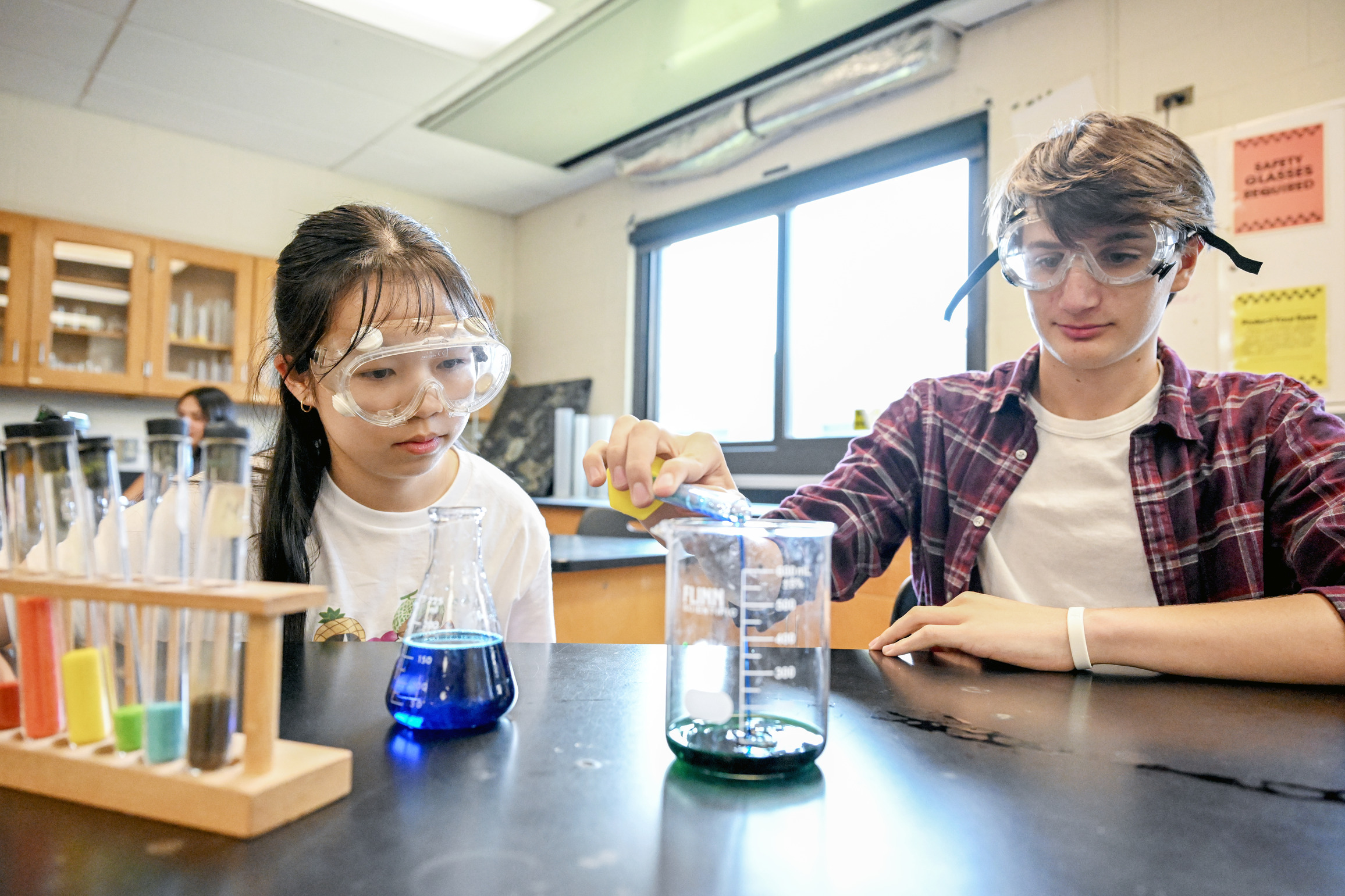 Two high school students wear protective goggles while pouring liquids into a beaker.