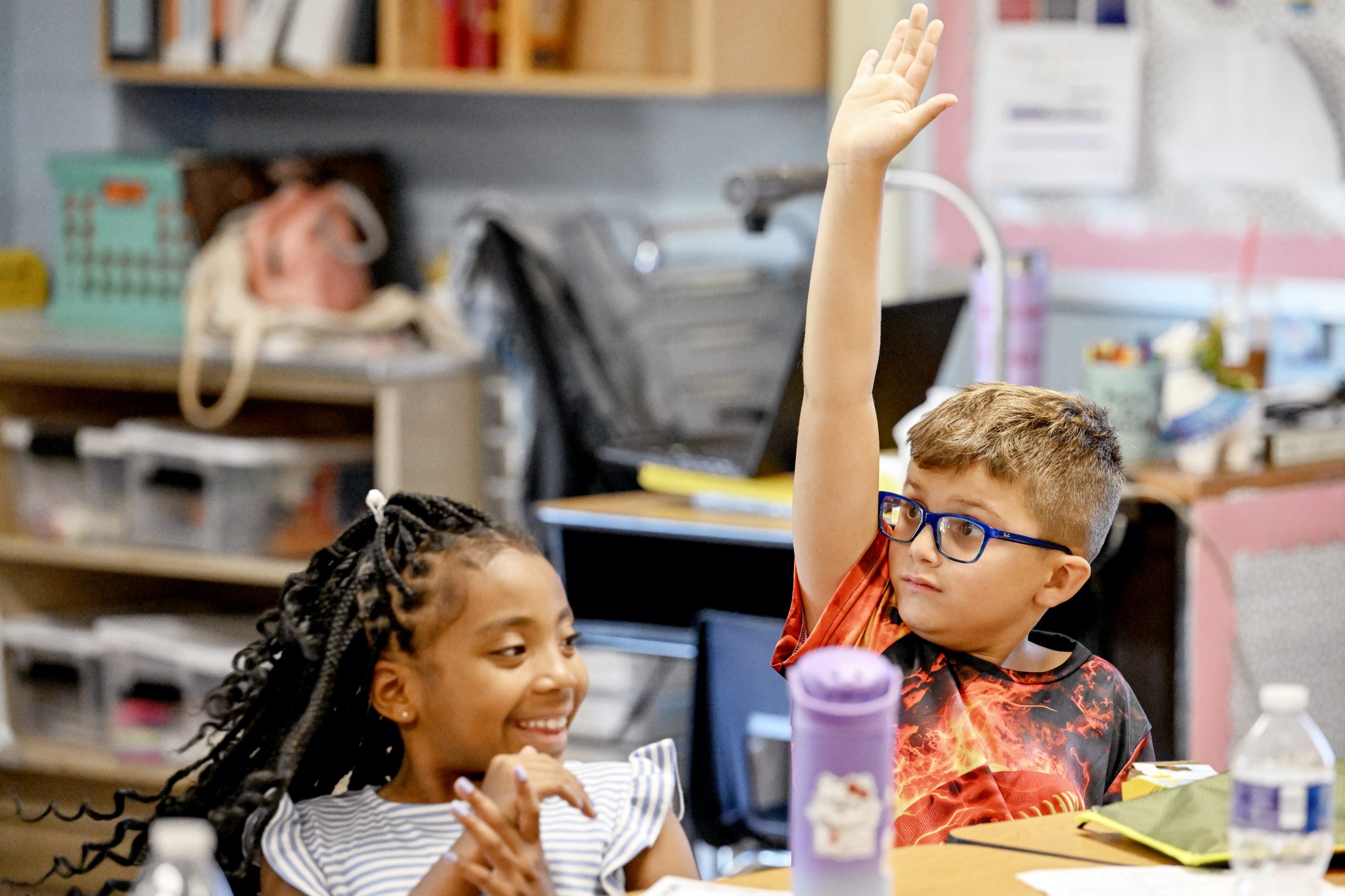 Young children, one with glasses and raising their hand, inside an elementary classroom