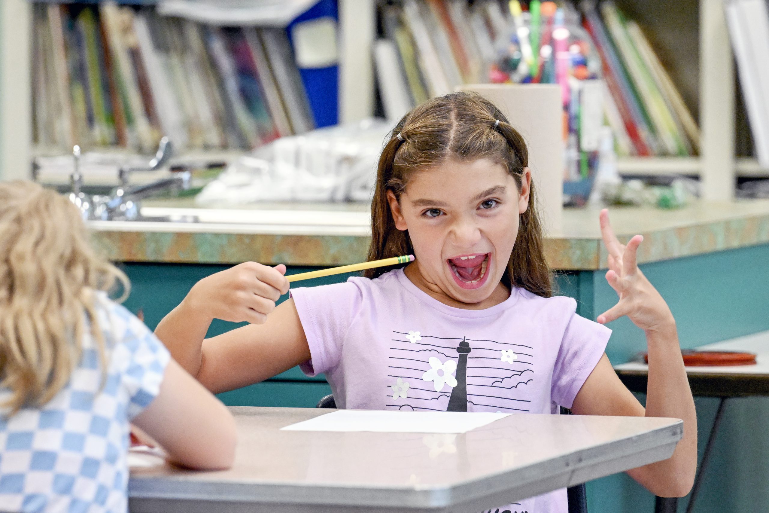 Young child grinning widely sitting at a desk in an elementary school classroom.